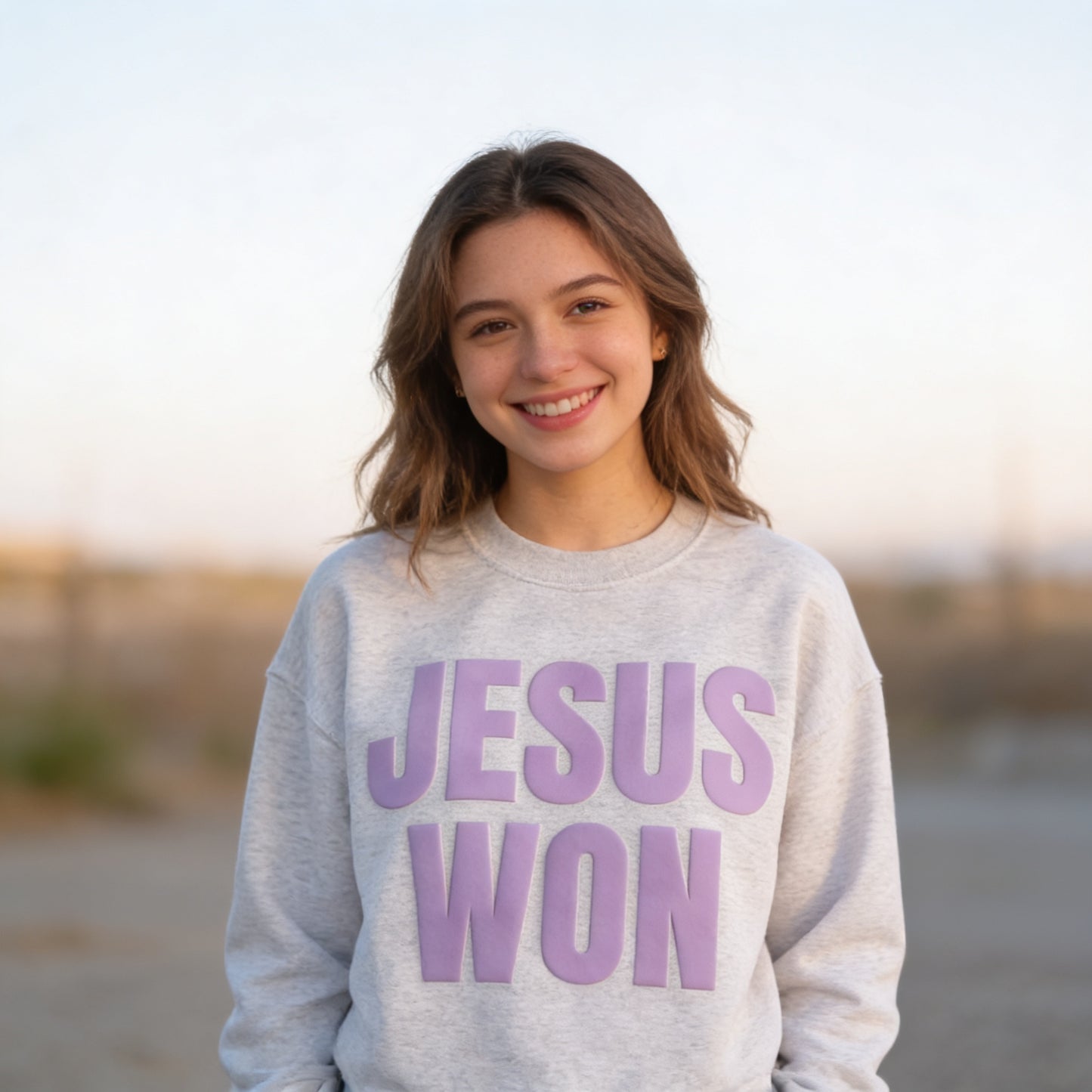 A young woman with long brown hair is smiling and wearing a grey sweatshirt with the text "JESUS WON" printed on it.