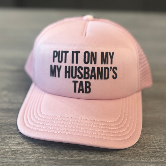 A pink baseball cap with the text "PUT IT ON MY HUSBAND'S TAB" printed on it.