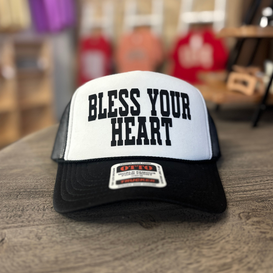 A white and black baseball cap with the text "Bless Your Heart" printed on it, placed on a wooden surface.