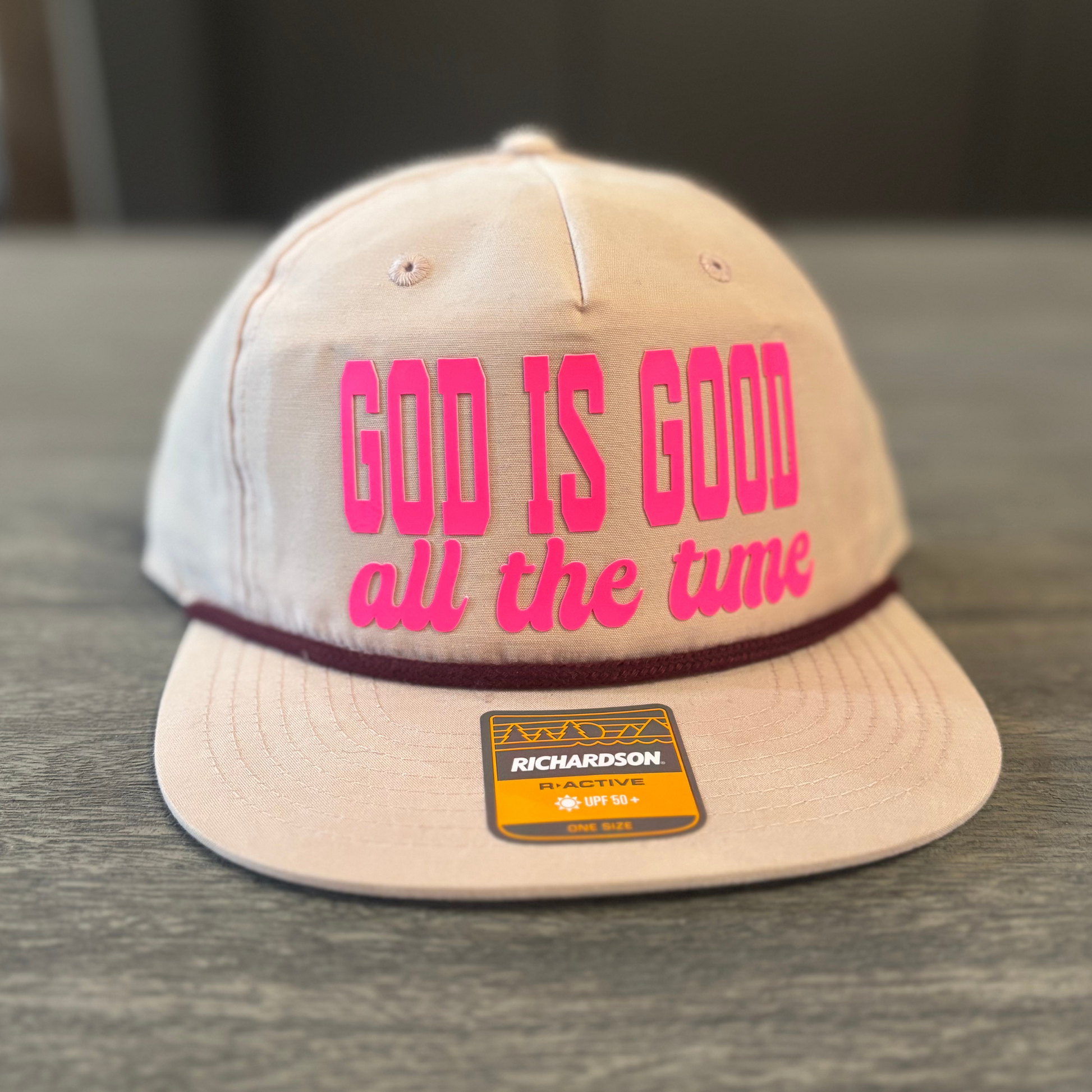 A beige baseball cap with the text "GOD IS GOOD all the time" printed on it in pink and orange letters.