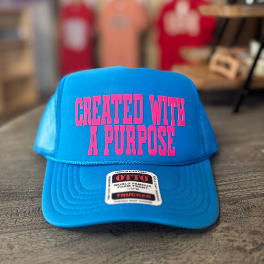 A blue baseball cap with the text "CREATED WITH A PURPOSE" printed on it, sitting on a wooden surface.