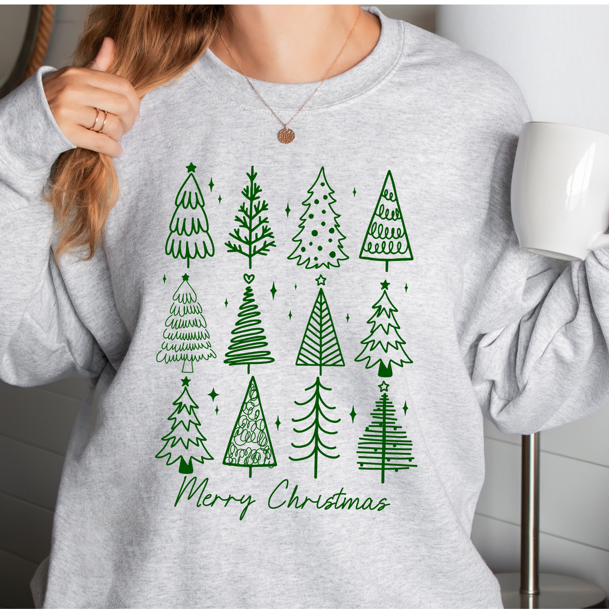 Woman wearing an ash sweatshirt with green doodle Christmas trees and Merry Christmas text.