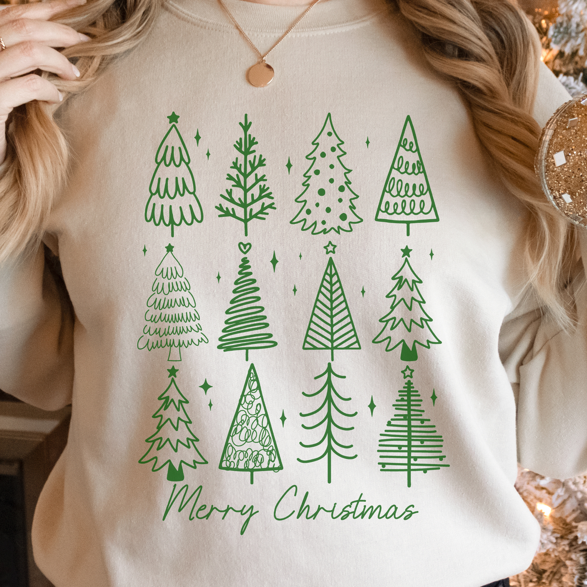 Woman wearing a sand sweatshirt with green doodle Christmas trees and Merry Christmas text.